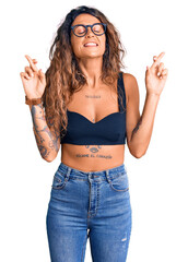 Young hispanic woman with tattoo wearing casual clothes and glasses gesturing finger crossed smiling with hope and eyes closed. luck and superstitious concept.