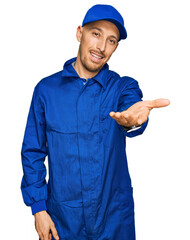 Bald man with beard wearing builder jumpsuit uniform smiling cheerful offering palm hand giving assistance and acceptance.