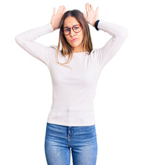 Beautiful brunette young woman wearing casual white sweater and glasses doing bunny ears gesture with hands palms looking cynical and skeptical. easter rabbit concept.
