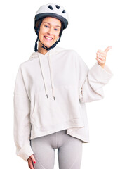 Beautiful brunette young woman wearing bike helmet and sporty clothes smiling with happy face looking and pointing to the side with thumb up.