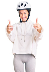 Beautiful brunette young woman wearing bike helmet and sporty clothes approving doing positive gesture with hand, thumbs up smiling and happy for success. winner gesture.