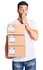 Young handsome hispanic man holding delivery package covering mouth with hand, shocked and afraid for mistake. surprised expression