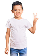 Little cute boy kid wearing casual white tshirt smiling with happy face winking at the camera doing victory sign. number two.