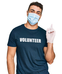 Hispanic young man wearing medical mask and volunteer t shirt smiling happy pointing with hand and finger to the side