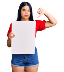 Young beautiful asian girl holding blank empty banner with angry face, negative sign showing dislike with thumbs down, rejection concept