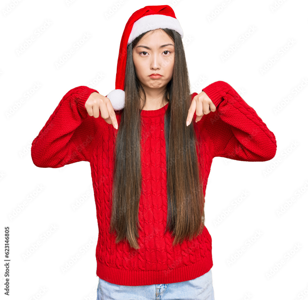 Sticker Young chinese woman wearing christmas hat pointing down looking sad and upset, indicating direction with fingers, unhappy and depressed.