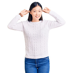 Young beautiful chinese woman wearing casual sweater smiling pulling ears with fingers, funny gesture. audition problem