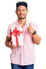 Handsome latin american young man holding gift smiling happy and positive, thumb up doing excellent and approval sign