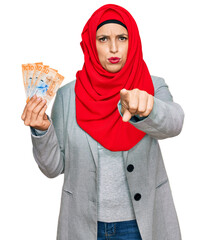 Beautiful hispanic woman wearing islamic hijab holding 10 swiss franc banknotes pointing with finger to the camera and to you, confident gesture looking serious