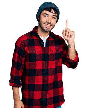 Hispanic young man with beard wearing wool winter hat smiling with an idea or question pointing finger up with happy face, number one