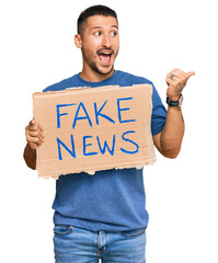 Handsome man with tattoos holding fake news banner pointing thumb up to the side smiling happy with open mouth