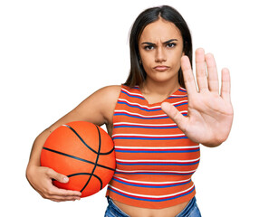 Young brunette woman holding basketball ball with open hand doing stop sign with serious and...