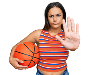 Young brunette woman holding basketball ball with open hand doing stop sign with serious and confident expression, defense gesture
