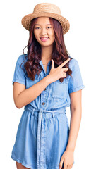 Young beautiful chinese girl wearing summer hat cheerful with a smile on face pointing with hand and finger up to the side with happy and natural expression