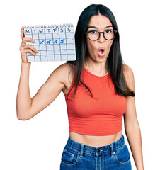 Young hispanic woman holding travel calendar scared and amazed with open mouth for surprise,...