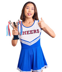 Youn beautiful asian girl wearing cheerleader uniform holding champion trophy smiling happy and positive, thumb up doing excellent and approval sign