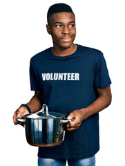 Young african american man wearing volunteer holding cooking pot smiling looking to the side and...