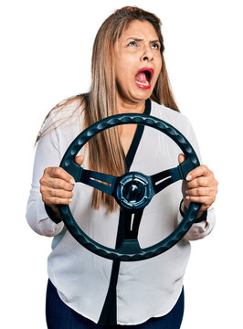 Middle age latin woman holding steering wheel angry and mad screaming frustrated and furious, shouting with anger looking up.