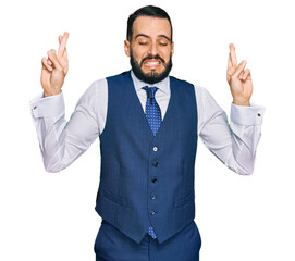 Young man with beard wearing business vest gesturing finger crossed smiling with hope and eyes closed. luck and superstitious concept.