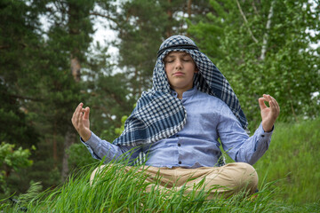 In summer, a boy in an Arab scarf sits in meditation on the grass.