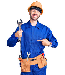 Young hispanic man wearing electrician uniform holding wrench pointing finger to one self smiling happy and proud
