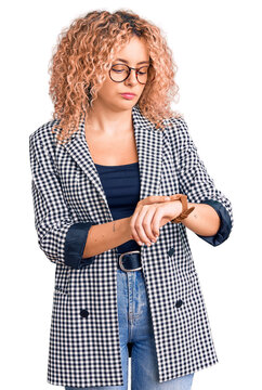 Young blonde woman with curly hair wearing business jacket and glasses checking the time on wrist watch, relaxed and confident
