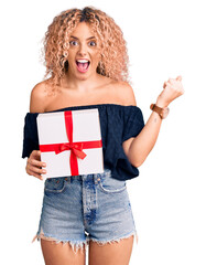 Young blonde woman with curly hair holding gift screaming proud, celebrating victory and success very excited with raised arms