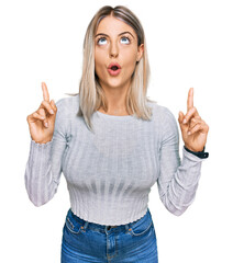 Beautiful blonde woman wearing casual clothes amazed and surprised looking up and pointing with fingers and raised arms.