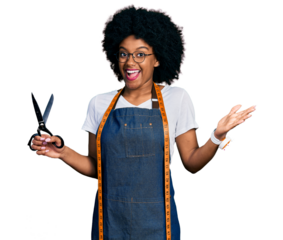 Young african american woman dressmaker designer wearing atelier apron holding scissors celebrating achievement with happy smile and winner expression with raised hand
