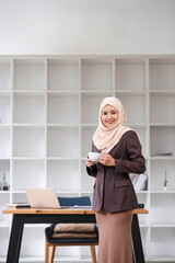 A portrait of a confident and smiling Asian Muslim businesswoman stands in her modern office with a coffee cup in her hands.