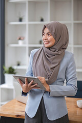 A portrait of a successful Asian Muslim businesswoman stands in her modern office with a digital...