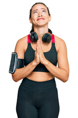 Beautiful blonde woman wearing gym clothes and using headphones begging and praying with hands together with hope expression on face very emotional and worried. begging.