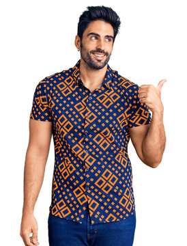 Handsome hispanic man wearing casual clothes smiling with happy face looking and pointing to the side with thumb up.