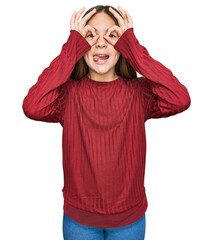 Beautiful brunette little girl wearing casual sweater doing ok gesture like binoculars sticking tongue out, eyes looking through fingers. crazy expression.