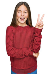 Beautiful brunette little girl wearing casual sweater smiling with happy face winking at the camera doing victory sign. number two.