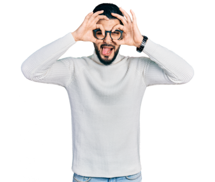 Young arab man with beard wearing elegant turtleneck sweater and glasses doing ok gesture like binoculars sticking tongue out, eyes looking through fingers. crazy expression.