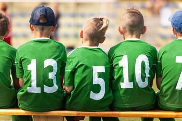 School soccer players sitting on wooden substitute bench. Group of children boys playing sports competition game. Kids in green sports uniforms