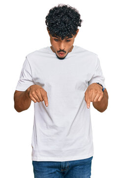 Young arab man wearing casual white t shirt pointing down with fingers showing advertisement, surprised face and open mouth