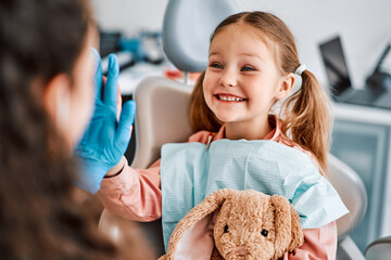 At the doctor's appointment. A candid emotional photo of a child sitting in a dental chair, holding a toy rabbit and cheerfully giving a high-five to the nurse. - obrazy, fototapety, plakaty