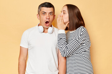 Shock, gossip, share advice. Young woman whispering to man on ear, brunette guy with open mouth excited and surprised, posing isolated over beige background