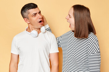 Negative emotion facial expression feelings, reaction, situation. Angry young woman pulling ear lobe of surprised shocked in pain hurting funny man, isolated over beige background.