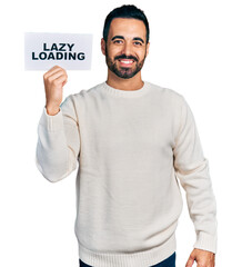 Young hispanic man with beard holding lazy loading banner looking positive and happy standing and smiling with a confident smile showing teeth