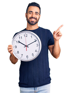 Young hispanic man holding big clock smiling happy pointing with hand and finger to the side
