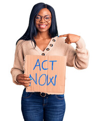 Young african american woman holding act now banner pointing finger to one self smiling happy and...