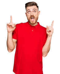 Young redhead man wearing casual clothes amazed and surprised looking up and pointing with fingers and raised arms.