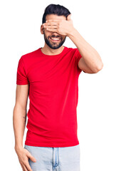 Young handsome man with beard wearing casual t-shirt smiling and laughing with hand on face covering eyes for surprise. blind concept.