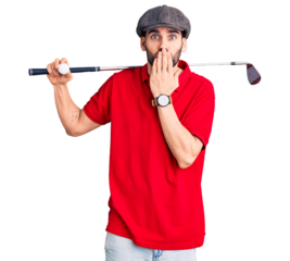 Young handsome man with beard playing golf holding club and ball covering mouth with hand, shocked and afraid for mistake. surprised expression