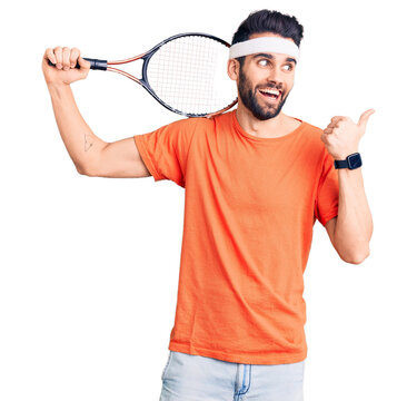 Young handsome man with beard playing tennis holding racket pointing thumb up to the side smiling happy with open mouth