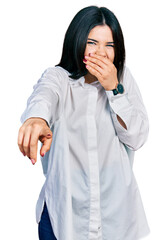 Young brunette woman with blue eyes wearing oversize white shirt laughing at you, pointing finger to the camera with hand over mouth, shame expression