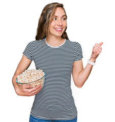 Young blonde woman eating popcorn pointing thumb up to the side smiling happy with open mouth